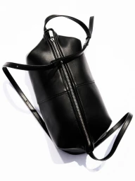 Black leather cylindrical barrel bag with central silver zipper and thin straps on a white background.