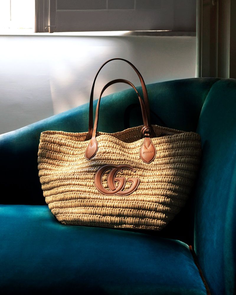 Gucci straw tote bag with brown leather trim and handles sitting on a blue velvet couch in a room with natural light.