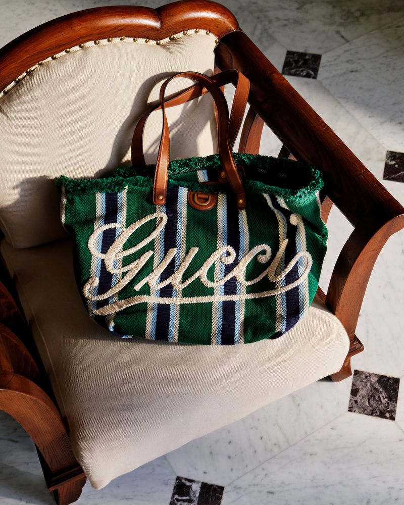 Green and navy striped Gucci tote bag with brown leather straps resting on a cream-colored cushioned chair in a well-lit indoor setting.