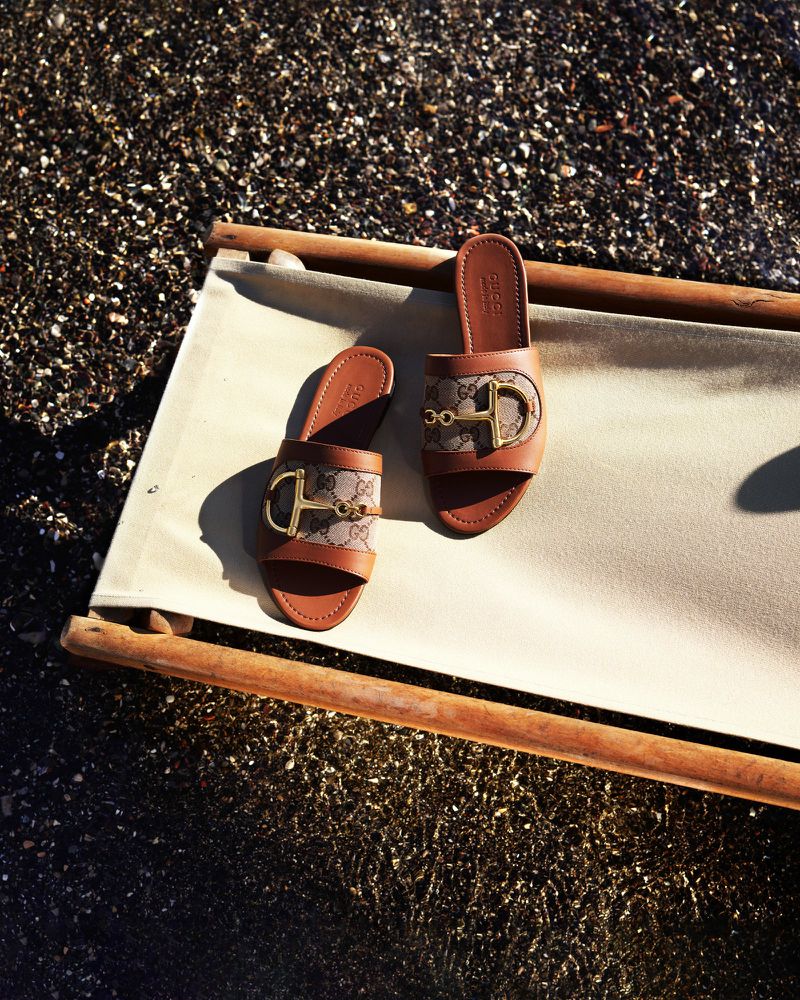 Gucci slide sandals with gold horsebit hardware displayed on a white beach chair against a dark sand beach background.
