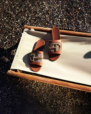 Pair of Gucci monogram horsebit slide sandals resting on a cream canvas beach chair over dark wet pebbles.