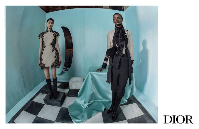 Two fashion models in modern attire pose in an aqua-colored studio against a checkered floor. The background features unique geometric details.