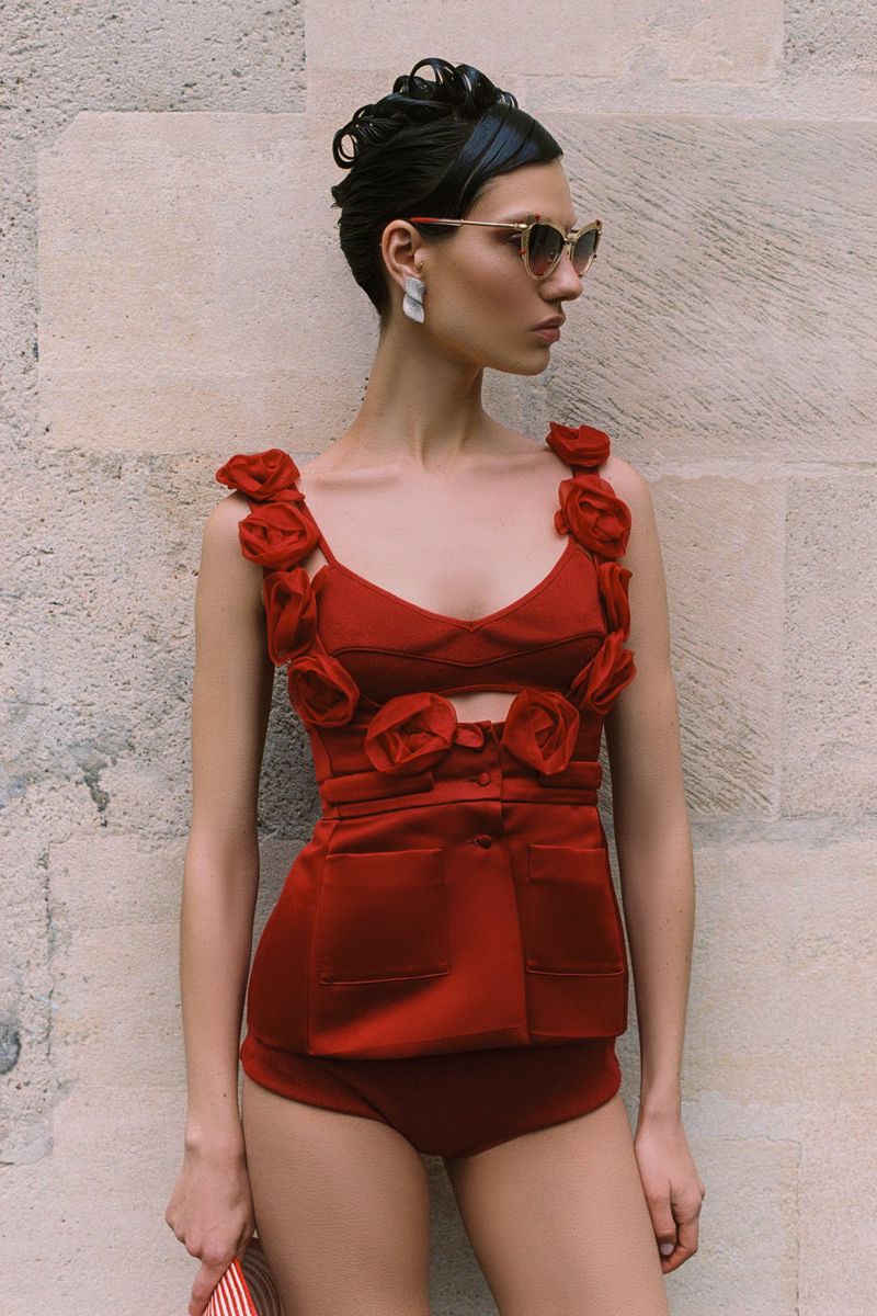 A female fashion model poses in a red two-piece outfit with rose details against a stone wall backdrop, showcasing a vintage-inspired style.