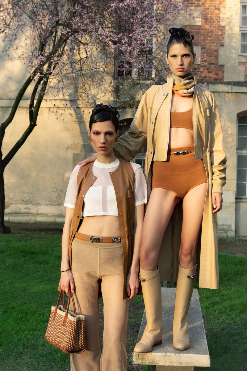 Two fashion models in matching outfits pose outside a historic building. One carries a woven bag, exuding high-fashion elegance.