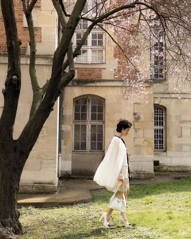 Adult female in a white dress and heels walks on the lawn near a tree with pink blossoms and a beige brick building with windows.