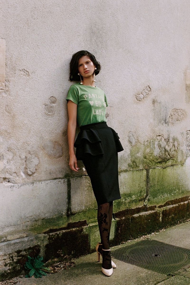 A fashionable woman stands in front of a textured wall wearing a green graphic tee, a black ruffled skirt, patterned tights, and heeled shoes.