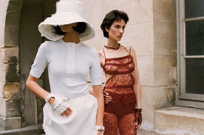 Two women pose outdoors in high fashion, one wearing white hat, top, skirt and gloves, the other wearing a brown lace dress.