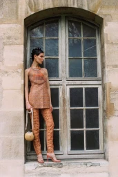 Model in patterned peach sleeveless tunic and trousers standing on an arched stone window ledge holding a round handbag.