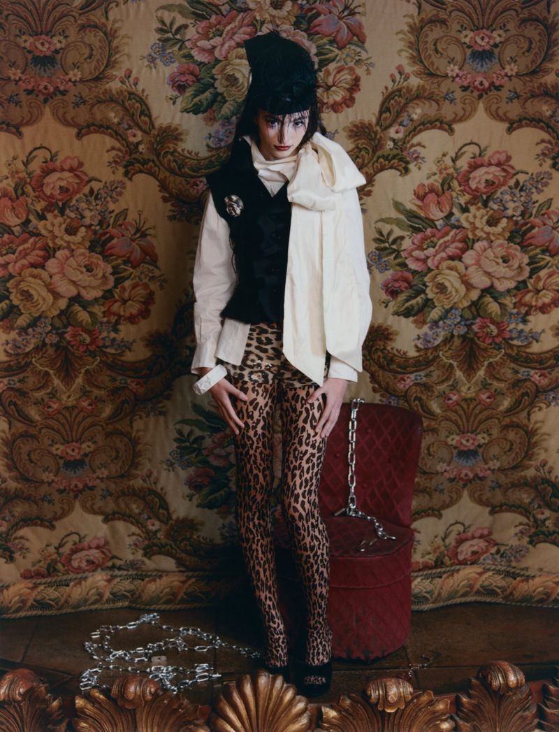 Model wearing leopard-print tights, a black vest, and a cream scarf standing before a floral tapestry with silver chains on the floor.
