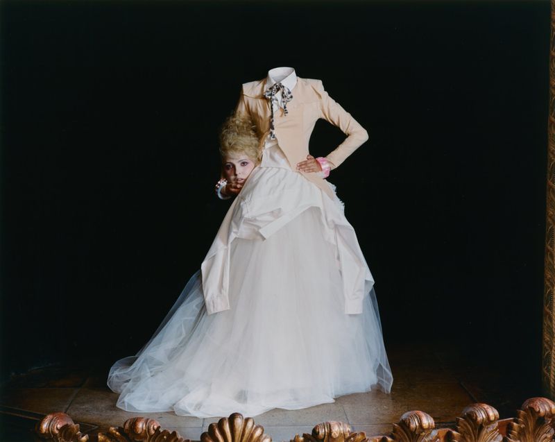 Surreal fashion portrait of a headless figure in a beige jacket and voluminous white tulle gown holding a woman's head underarm.