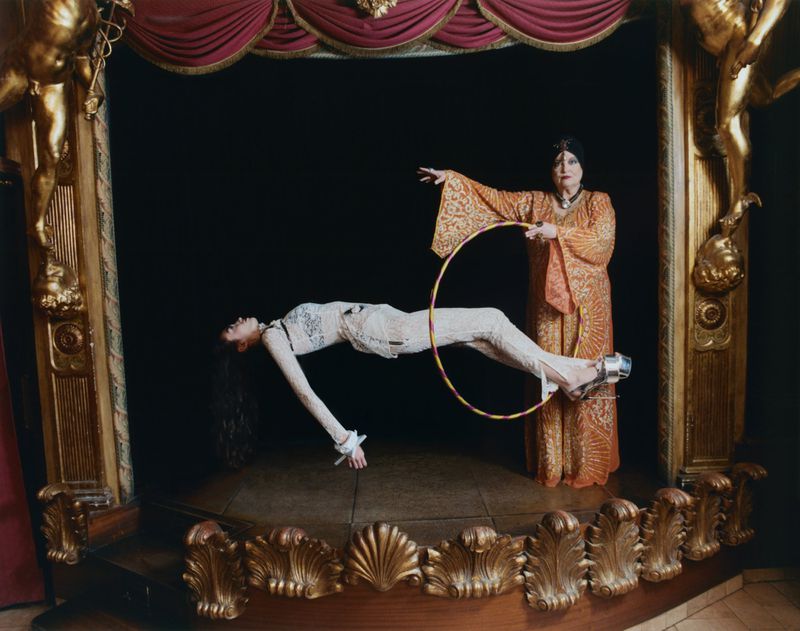 A woman in a cream jumpsuit appears to levitate horizontally inside an ornate golden stage as another woman holds a hoop.