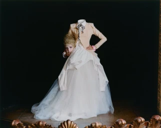 Surreal fashion portrait of a headless figure in a beige jacket and voluminous white tulle gown holding a woman's head underarm.