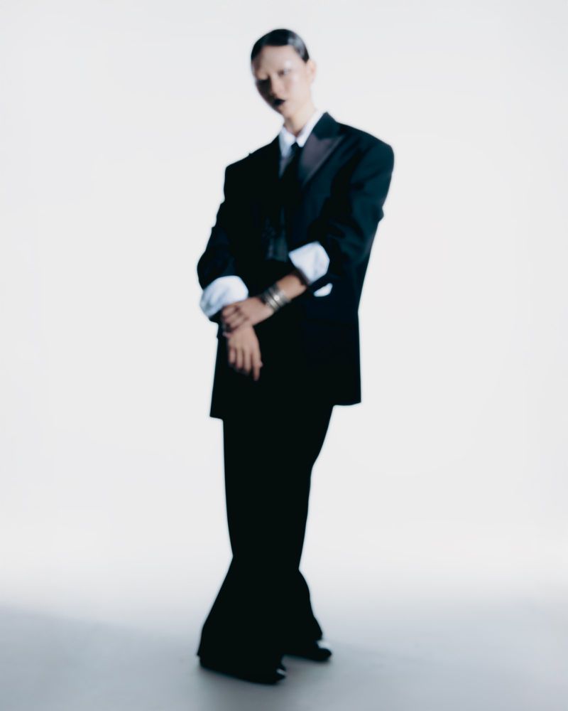 A person in a black suit standing in a photo studio with arms crossed, wearing bracelets and dark lipstick, against a white backdrop.