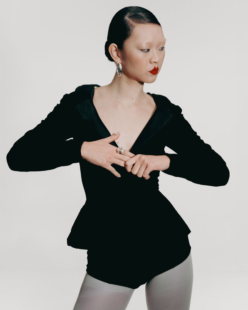 An Asian woman is posed in a photo studio wearing a black peplum top with long sleeves, light grey tights, red lipstick, and silver earrings.