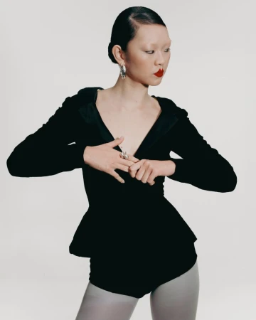 An Asian woman is posed in a photo studio wearing a black peplum top with long sleeves, light grey tights, red lipstick, and silver earrings.