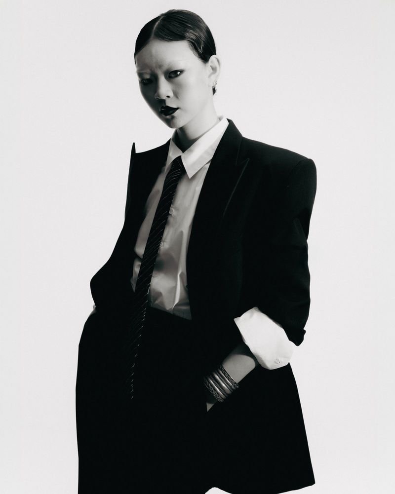 Monochrome portrait of a woman in a dark suit with hands in pockets, standing in a studio with a blank white background.