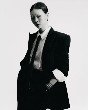 Monochrome portrait of a woman in a dark suit with hands in pockets, standing in a studio with a blank white background.