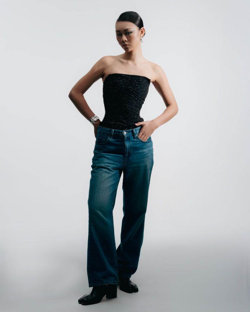 Portrait of a young Asian woman in a black tube top and denim jeans against a white backdrop in a fashion photography style.