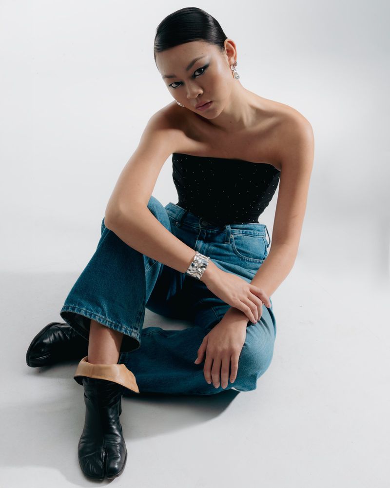 A stylish young Asian woman sits in a photo studio wearing a black bustier top, wide blue jeans, statement silver cuff, and black tabi boots.