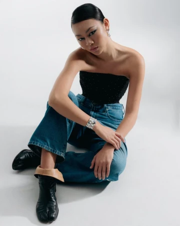 A stylish young Asian woman sits in a photo studio wearing a black bustier top, wide blue jeans, statement silver cuff, and black tabi boots.