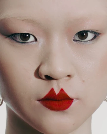Closeup portrait of an Asian woman with red geometric lipstick, dramatic eyeliner and pale complexion against a pale background. 