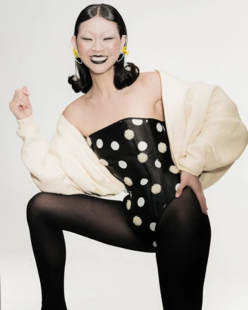 Stylish young woman in polka dot bodysuit and black tights posing against a white backdrop with a soft yellow jacket.