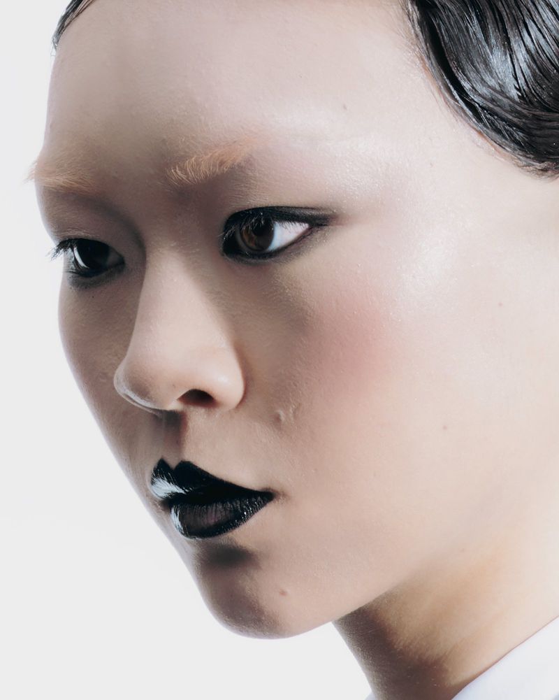 Close-up portrait of an adult woman with wet-look hair, wearing black lipstick, and makeup against a bright white backdrop.