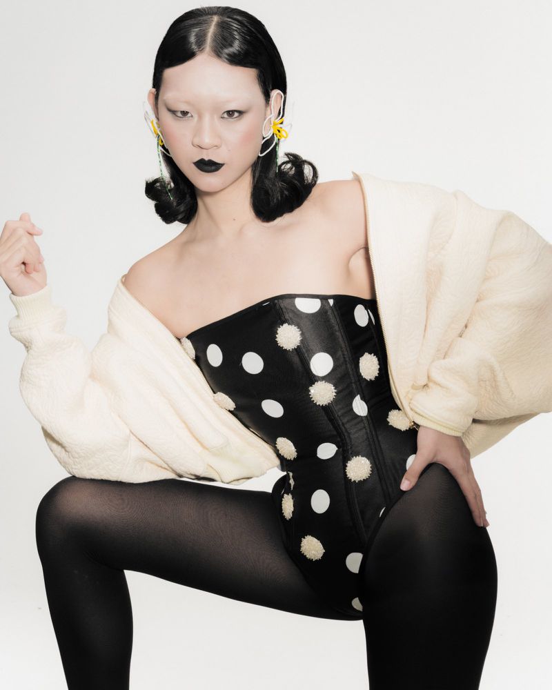 Portrait of a woman wearing a polka dot corset and black tights, with an off-white jacket draped over her shoulders, set against a white backdrop.