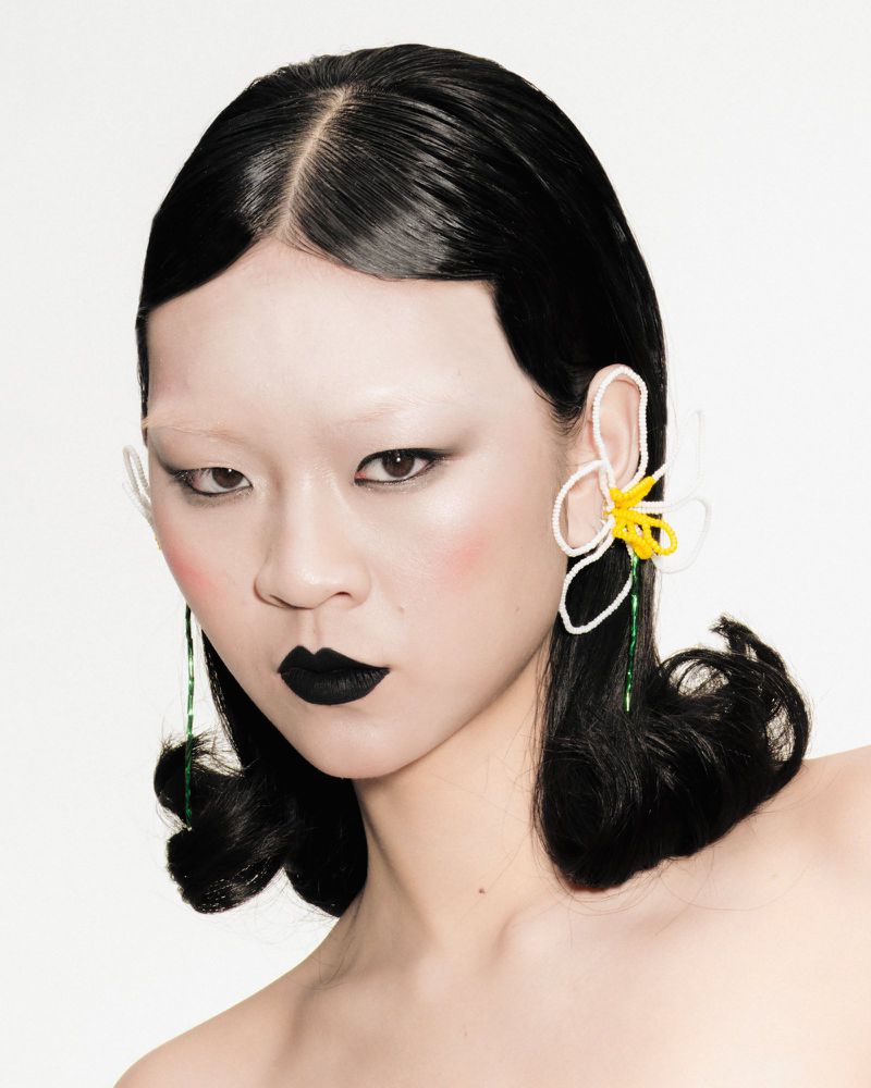 Portrait of a person with dark hair and flower earrings. The subject has pale skin, dark lipstick, and is posing in front of a white backdrop.