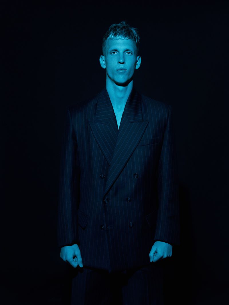Man wearing a dark pinstripe double-breasted suit illuminated in blue light against a dark background