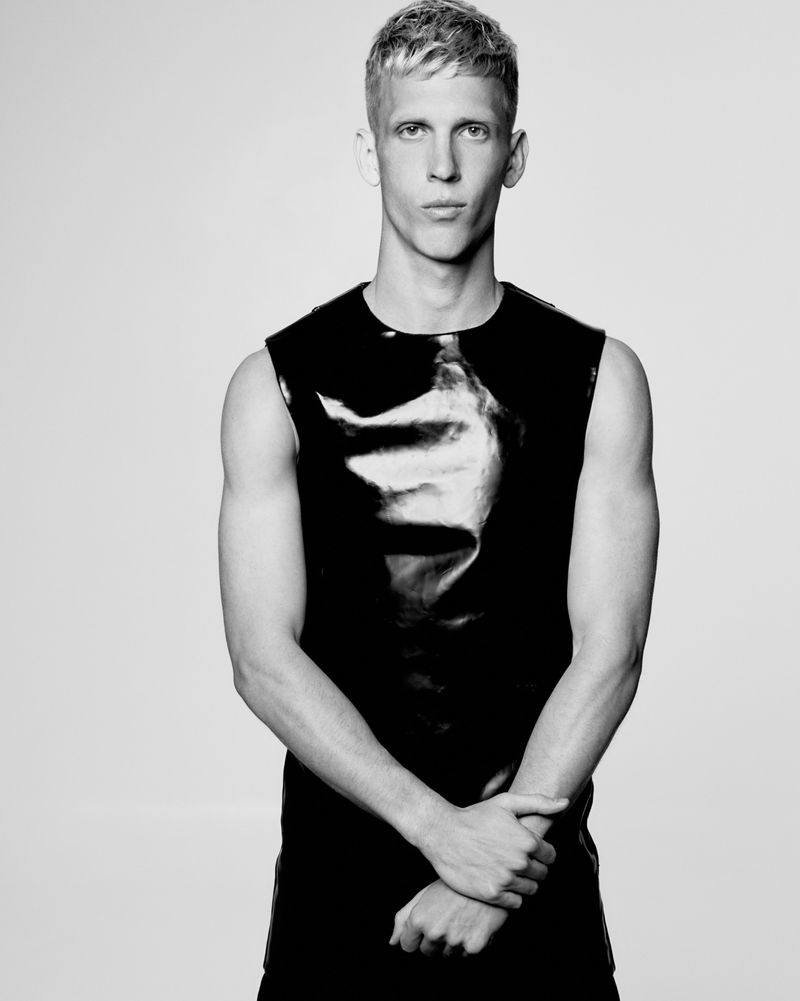 Dani Olmo posing in a black sleeveless glossy top with hands clasped against a light studio backdrop in black and white.
