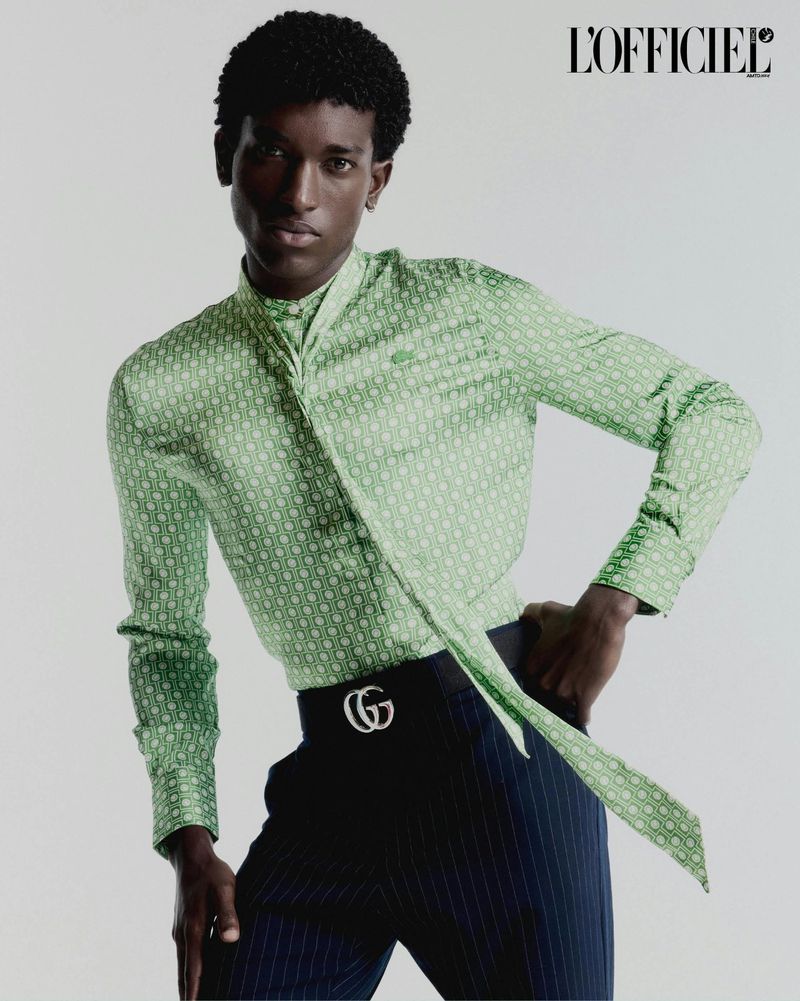 Gabriel Pitta poses in a patterned green long-sleeve shirt with neck tie and navy pinstripe trousers against a pale studio backdrop.