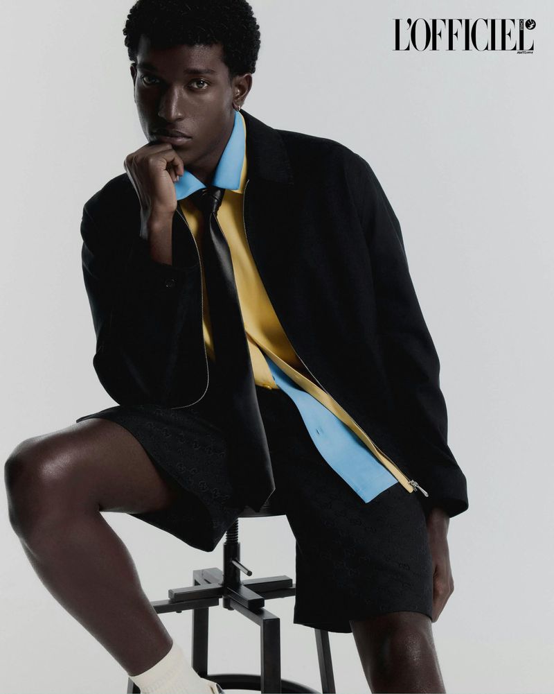 Model Gabriel Pitta seated on a studio stool wearing a black jacket, tie, yellow shirt, and black shorts on a white backdrop.