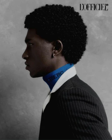 Profile portrait of a Black man with textured afro hair wearing a blue turtleneck and pinstripe blazer against a grey backdrop.