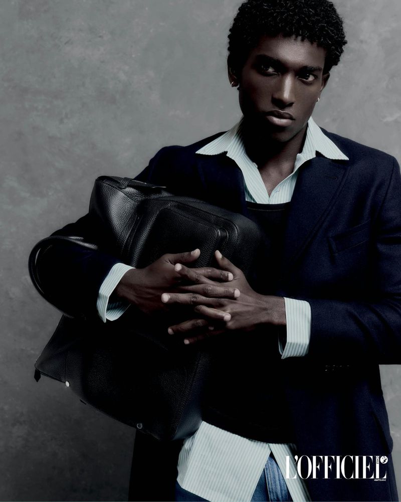 Model Gabriel Pitta holding a black leather tote bag, wearing a dark navy blazer over a light striped shirt against a grey backdrop.