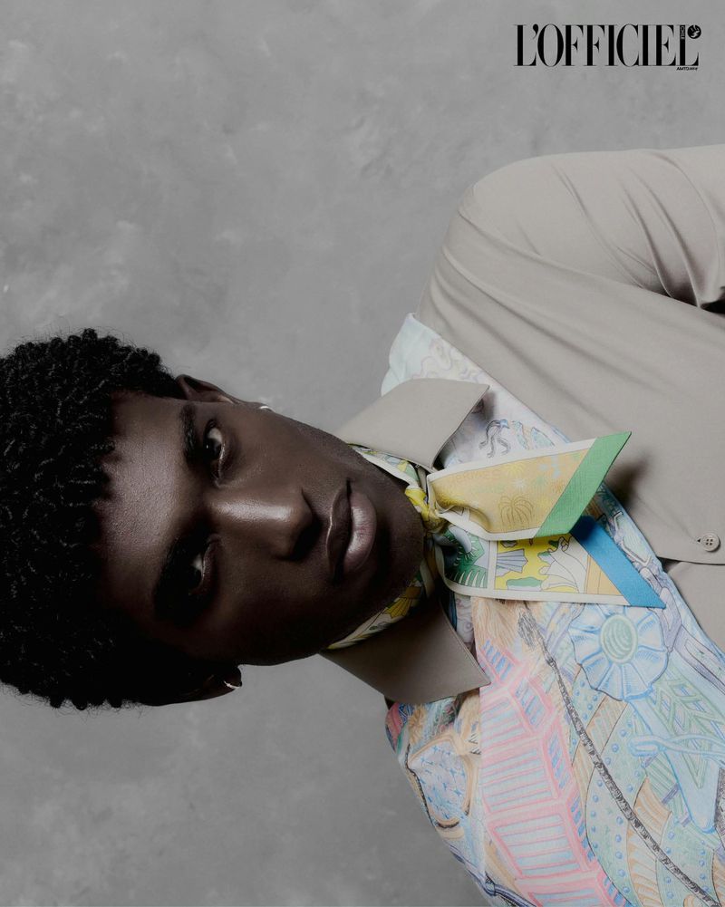 Gabriel Pitta wearing a beige collared shirt and a pastel patterned scarf against a textured grey studio backdrop.