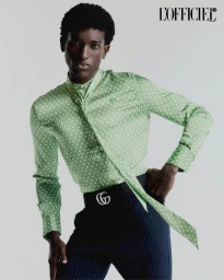 Gabriel Pitta poses in a patterned green long-sleeve shirt with neck tie and navy pinstripe trousers against a pale studio backdrop.