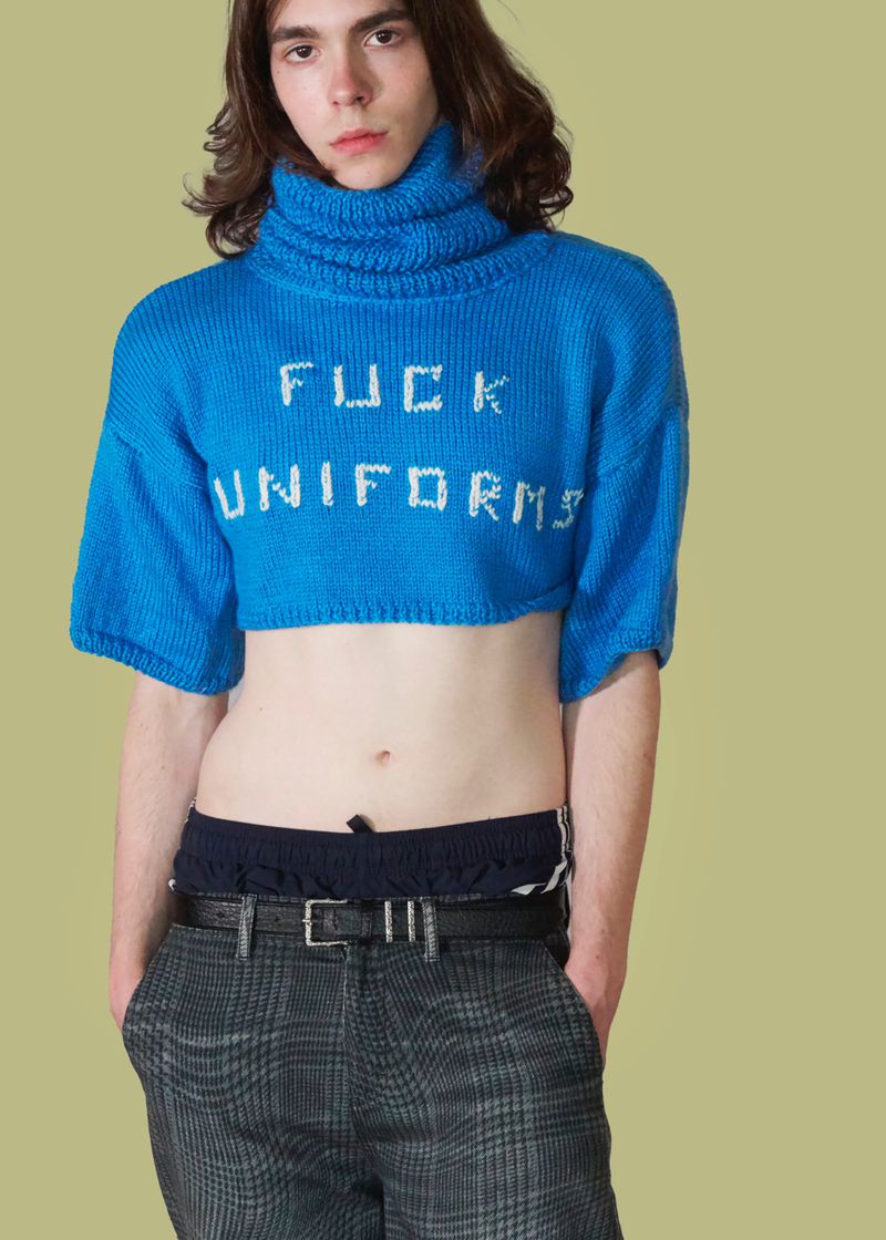 Model wearing a cropped blue turtleneck knit sweater with white text and plaid trousers against a pale green backdrop.