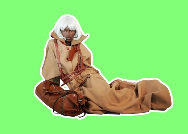 Model Adhieu Arok in a white bob wig and oversized tan coat with leather harness straps reclining against a bright green background.