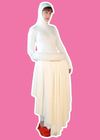 Model posing in a sculptural white hooded ensemble with a draped asymmetrical skirt and red shoes against a bright pink background.