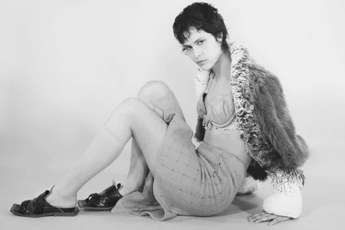 Model with short dark hair seated on the floor wearing a textured skirt, bralette, and fur jacket on a white seamless backdrop.
