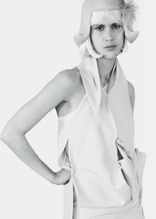 Model wearing a pale draped halter top and a vintage-style bob wig with a feather accent against a pure white background.