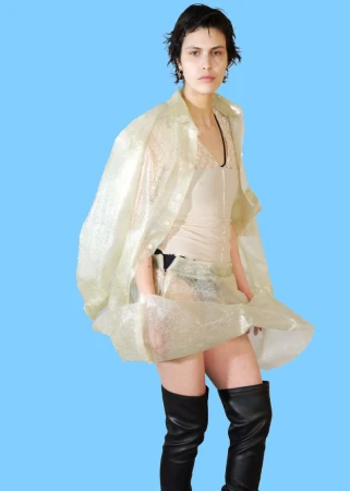 Model wearing a sheer iridescent wrap coat and black thigh-high leather boots against a solid sky blue studio backdrop.