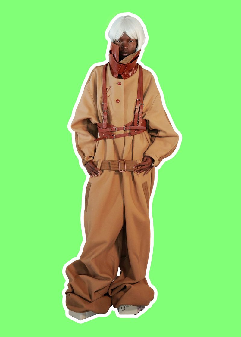 Model Adhieu Arok wearing an oversized camel jumpsuit with brown leather harness detailing against a solid green background.