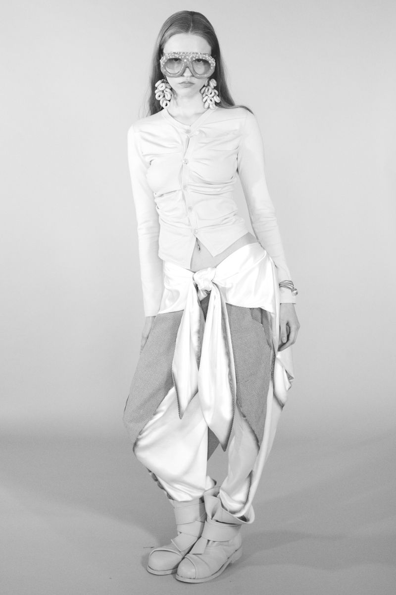 Model wearing statement goggles, earrings, patterned top, layered pants and boots against a plain grey seamless background.