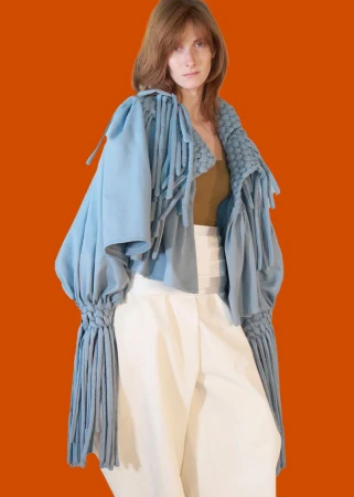 Model wearing an avant-garde powder blue fringed jacket and high-waisted white trousers against a solid orange background.