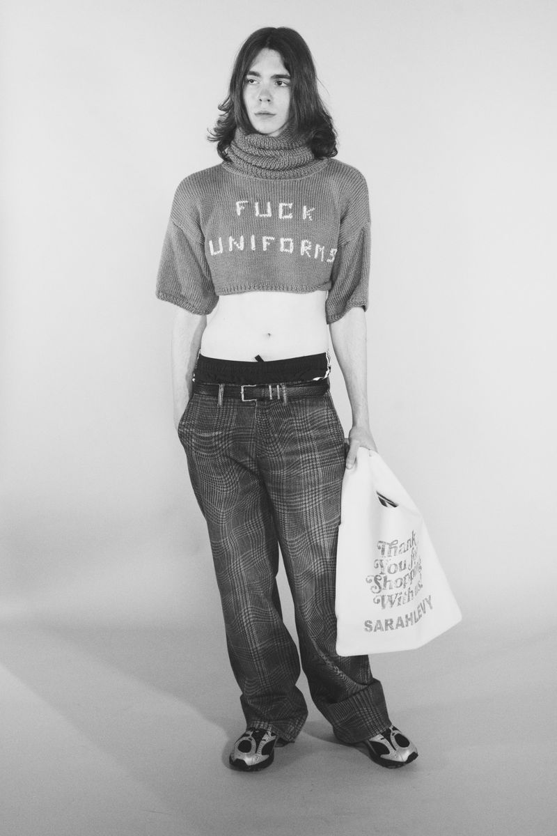 Model wearing a cropped turtleneck sweater reading Fuck Uniforms and plaid trousers holding a tote bag in a studio portrait.