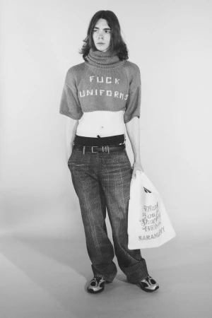 Model wearing a cropped turtleneck sweater reading Fuck Uniforms and plaid trousers holding a tote bag in a studio portrait.