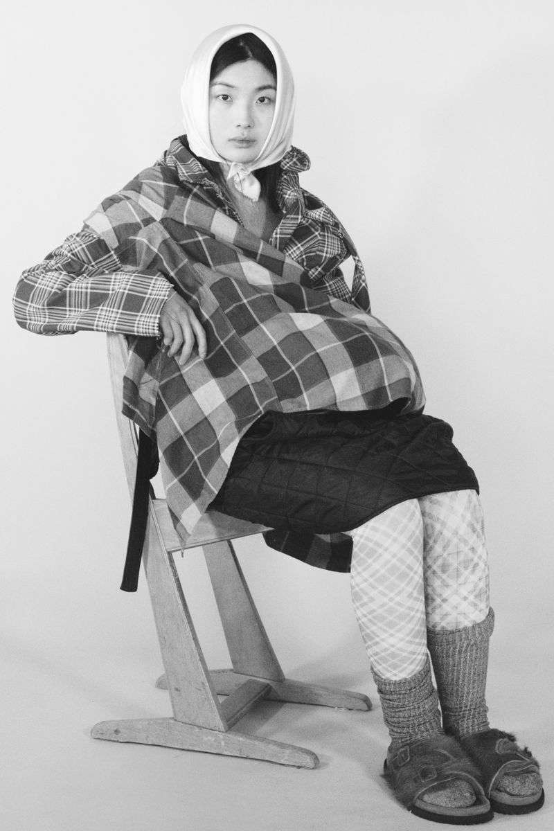 Monochrome fashion portrait of an East Asian woman seated on a wooden chair wearing an oversized plaid jacket and headscarf.