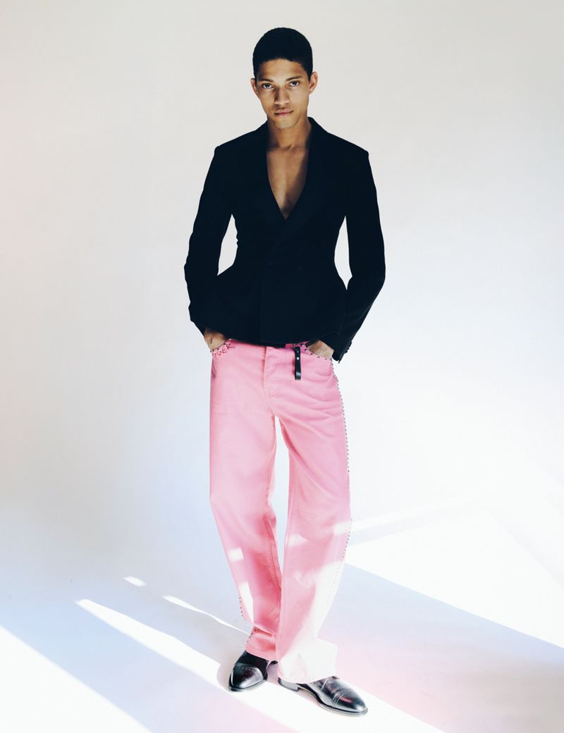 Full-length portrait of a male model wearing a black blazer and pink trousers against a seamless white studio background.